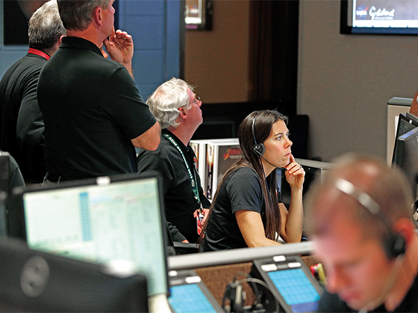 A  female student sits in a workplace setting with computers and screens. A group of male professionals are situated around her in the room. All are wearing lanyards and headsets and watching screens or monitors.